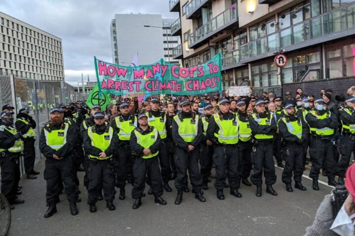 Glasgow, 3 novembre 2021. La polizia blocca la marcia di Extinction Rebellion che si dirigeva nei paraggi della Cop26. Lo striscione gioca sul doppio senso della parola "cop", in inglese poliziotto: quante cop (e quanti "cops") ci vogliono per arrestare il cambiamento climatico?