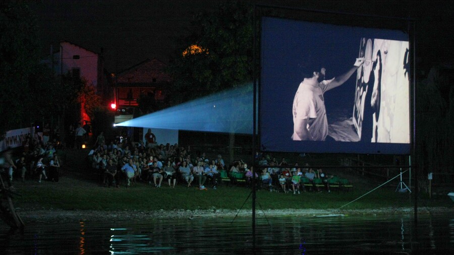 Sullo schermo montato dentro il lago si proiettano le immagini di un film in bianco e nero. Sulla riva il pubblico assiste seduto su sedie sul prato