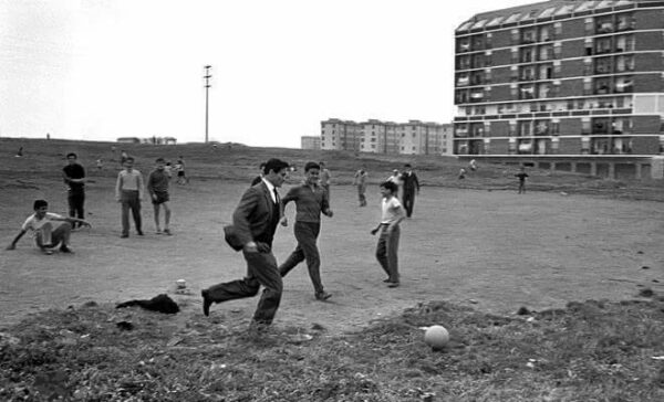 Pasolini gioca a pallone con dei bambini, sullo sfondo palazzoni moderni nella periferia romana